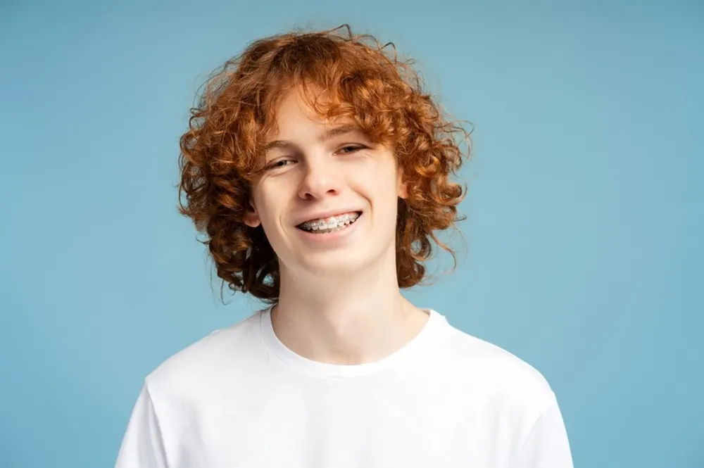 A teenage boy with red curly hair and fixed braces smiling against a blue background