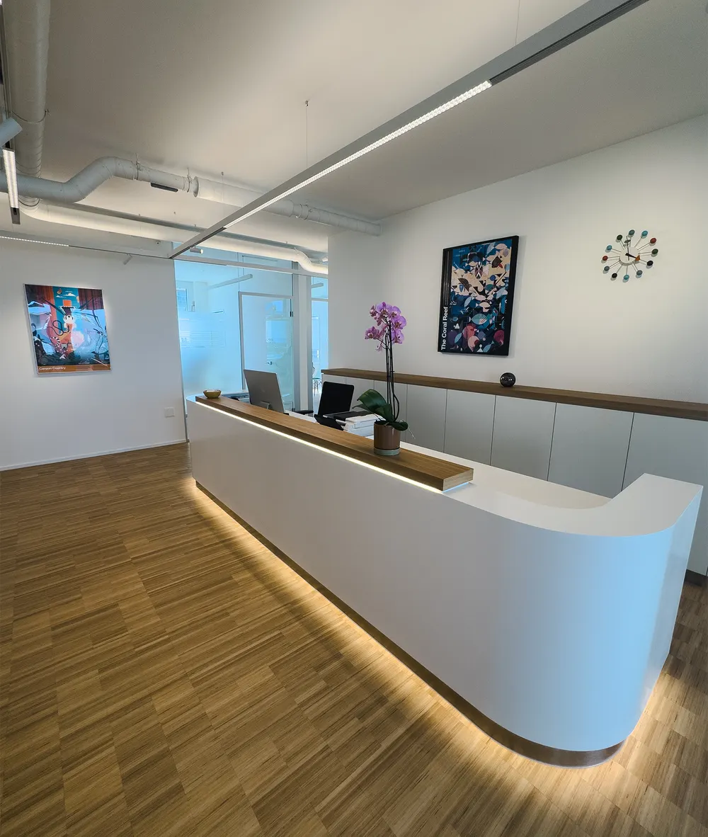 Reception desk with a white counter and wooden top, the workstation behind it and a glass wall to the treatment area