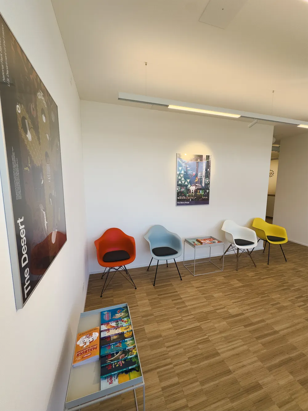 Waiting area with coloured shell chairs, two art posters on the wall and a table of children’s books